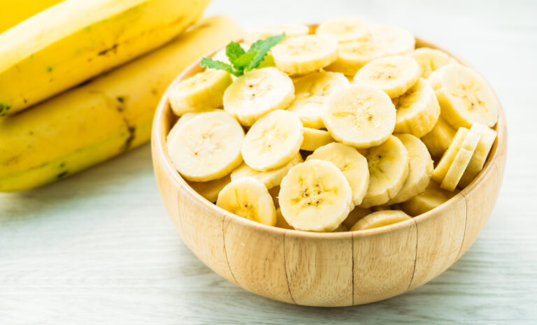 Bananas frescas e fatiadas em uma tigela de madeira, com bananas inteiras ao fundo. A banana é uma opção nutritiva e versátil para substituir açúcar em vitaminas, bolos e doces naturais.