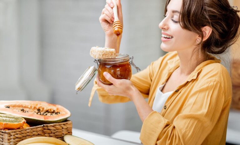 Mulher sorridente, vestindo camisa amarela, despejando mel de um pote de vidro. O mel é um adoçante natural que pode substituir açúcar em diversas preparações, trazendo sabor e benefícios nutricionais.