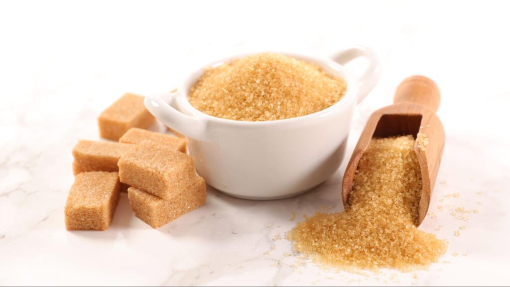 Tigela branca com açúcar mascavo granulado e cubos ao redor, com uma colher de madeira espalhando o açúcar. O açúcar mascavo pode substituir açúcar refinado, oferecendo um sabor mais intenso e nutrientes adicionais.